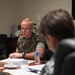 A U.S. Marine in MARPAT camouflage uniform is seated at a conference table, appearing to speak during a formal meeting. A name placard in Japanese script is visible on the table in front of him, along with documents, a water bottle, and a tea cup. A second individual in the foreground, partially visible and out of focus, appears to be wearing a camouflage uniform as well.