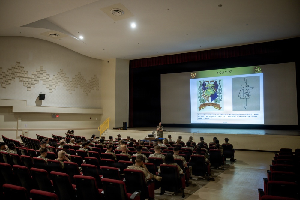 A military briefing or lecture is taking place in an auditorium-style theater, with a presenter standing at the front near a large projection screen. The audience consists of uniformed military personnel seated in red theater chairs. The presentation slide displays the date '4 Oct 1927' along with military insignia and a historical quote.