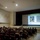 A military briefing or lecture is taking place in an auditorium-style theater, with a presenter standing at the front near a large projection screen. The audience consists of uniformed military personnel seated in red theater chairs. The presentation slide displays the date '4 Oct 1927' along with military insignia and a historical quote.