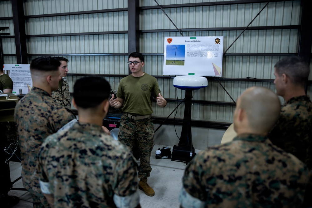 A U.S. Marine in an olive drab t-shirt is conducting a briefing or demonstration for a group of Marines in MARPAT camouflage uniforms inside what appears to be a hangar or warehouse facility. The presenter stands in front of a display board titled 'Sensors and Collectors' featuring two unit insignia patches and imagery of what appears to be radar or sensor equipment. A white dome-shaped sensor or antenna device is mounted on a stand beside the presenter.