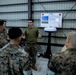 A U.S. Marine in an olive drab t-shirt is conducting a briefing or demonstration for a group of Marines in MARPAT camouflage uniforms inside what appears to be a hangar or warehouse facility. The presenter stands in front of a display board titled 'Sensors and Collectors' featuring two unit insignia patches and imagery of what appears to be radar or sensor equipment. A white dome-shaped sensor or antenna device is mounted on a stand beside the presenter.