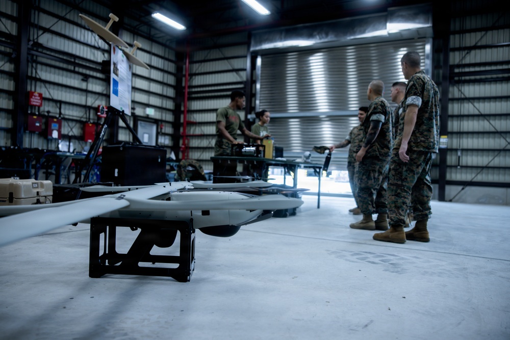 Military personnel in camouflage uniforms are gathered inside a hangar facility with unmanned aerial vehicles (UAVs/drones). A fixed-wing drone on a ground support stand is prominently visible in the foreground, while service members appear to be conducting training or maintenance operations. The setting appears to be a military UAV operations or maintenance facility.