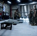 Military personnel in camouflage uniforms are gathered inside a hangar facility with unmanned aerial vehicles (UAVs/drones). A fixed-wing drone on a ground support stand is prominently visible in the foreground, while service members appear to be conducting training or maintenance operations. The setting appears to be a military UAV operations or maintenance facility.