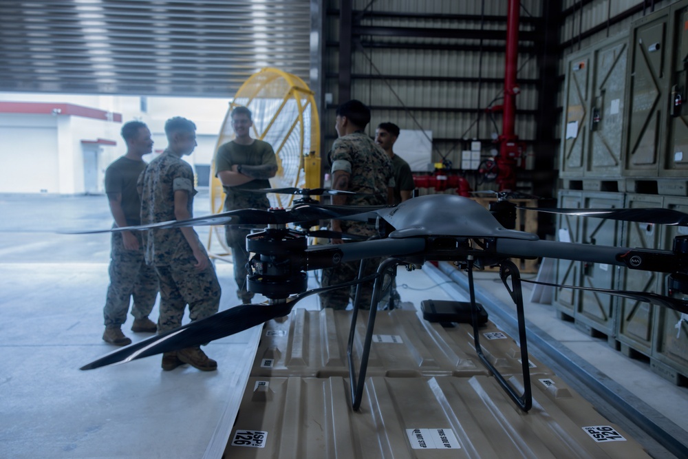 A military drone (quadcopter/multirotor UAS) is positioned in the foreground on what appears to be cardboard or packaging material inside a hangar or warehouse facility. Several personnel in military camouflage uniforms are gathered in the background, appearing to discuss or inspect equipment. The setting suggests a military logistics, training, or equipment familiarization context.