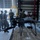 A military drone (quadcopter/multirotor UAS) is positioned in the foreground on what appears to be cardboard or packaging material inside a hangar or warehouse facility. Several personnel in military camouflage uniforms are gathered in the background, appearing to discuss or inspect equipment. The setting suggests a military logistics, training, or equipment familiarization context.