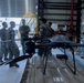 A military drone (quadcopter/multirotor UAS) is positioned in the foreground on what appears to be cardboard or packaging material inside a hangar or warehouse facility. Several personnel in military camouflage uniforms are gathered in the background, appearing to discuss or inspect equipment. The setting suggests a military logistics, training, or equipment familiarization context.