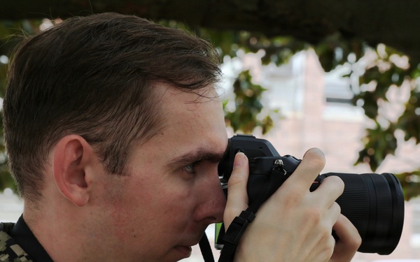 A young male in U.S. Navy Working Uniform (NWU Type III, woodland digital camouflage) is photographing with a DSLR camera raised to his eye. He is positioned outdoors beneath a tree, with a brick building and a parked dark-colored SUV visible in the background. The image appears to depict a military photographer or documentation activity.