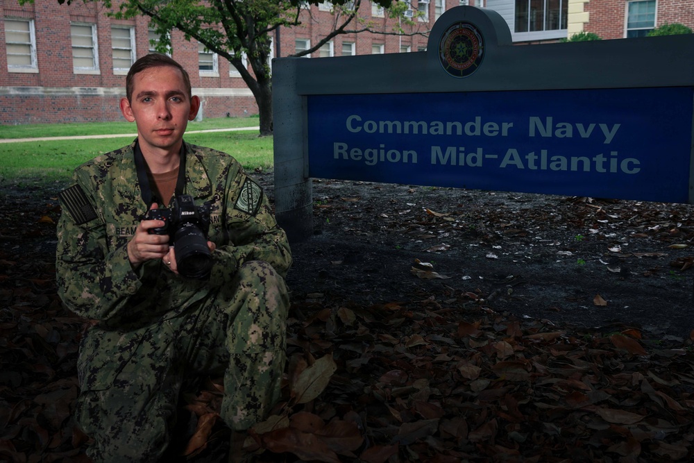 A U.S. Navy service member in NWU Type III (digital camouflage) uniform is posed kneeling in front of the 'Commander Navy Region Mid-Atlantic' sign. The individual is holding a Nikon DSLR camera with a lens attached, suggesting a role as a military photographer or Mass Communication Specialist. The setting appears to be a naval installation, likely in the Mid-Atlantic region of the United States.