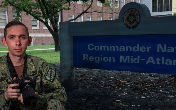 A U.S. Navy service member in NWU Type III (digital camouflage) uniform is posed kneeling in front of the 'Commander Navy Region Mid-Atlantic' sign. The individual is holding a Nikon DSLR camera with a lens attached, suggesting a role as a military photographer or Mass Communication Specialist. The setting appears to be a naval installation, likely in the Mid-Atlantic region of the United States.
