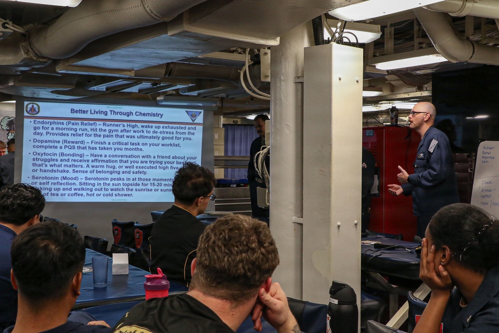 A military briefing or training session is taking place aboard what appears to be a naval vessel, with a presenter standing near a projected slide titled 'Better Living Through Chemistry.' The presentation covers neurochemicals including Endorphins, Dopamine, Oxytocin, and Serotonin and their roles in well-being. Several uniformed and civilian personnel are seated at tables facing the presenter and projection screen.
