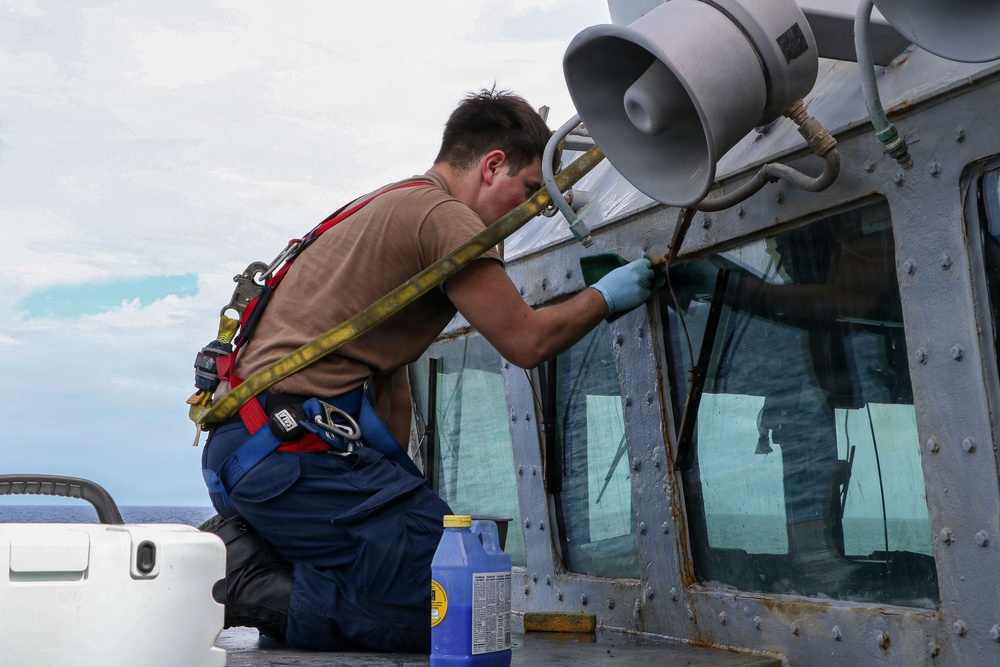 A sailor or maintenance worker wearing a safety harness is kneeling on the exterior deck of a naval vessel, performing maintenance work on the bridge windows. The individual is wearing blue gloves and working with a tool on the window frames, which show signs of rust and wear. Cleaning supplies, including a blue bottle of liquid, are visible nearby.