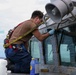 A sailor or maintenance worker wearing a safety harness is kneeling on the exterior deck of a naval vessel, performing maintenance work on the bridge windows. The individual is wearing blue gloves and working with a tool on the window frames, which show signs of rust and wear. Cleaning supplies, including a blue bottle of liquid, are visible nearby.