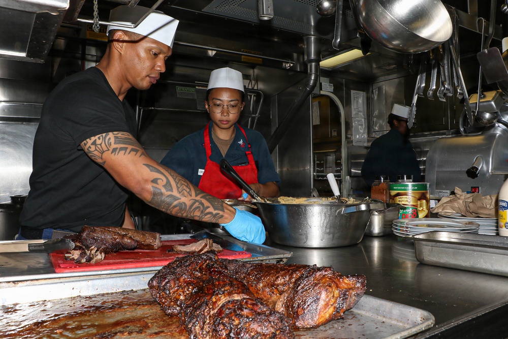 Two primary kitchen workers are preparing food in a professional stainless steel kitchen, with a third person visible in the background. Large roasted or smoked meat pieces are prominently displayed on the counter in the foreground. The workers are wearing chef's whites/hats and aprons, suggesting a formal food service environment.