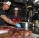 Two primary kitchen workers are preparing food in a professional stainless steel kitchen, with a third person visible in the background. Large roasted or smoked meat pieces are prominently displayed on the counter in the foreground. The workers are wearing chef's whites/hats and aprons, suggesting a formal food service environment.