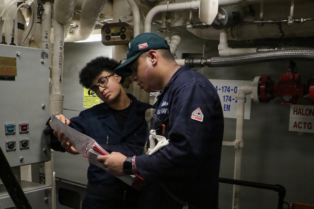 Two U.S. Navy personnel are reviewing documents together in what appears to be a ship's engineering or mechanical space. One individual wears a dark navy cap with an American flag patch and a 'U.S. Navy' labeled uniform, while the other wears glasses and a dark coverall. They appear to be conducting a technical review or inspection using printed documentation.