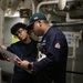 Two U.S. Navy personnel are reviewing documents together in what appears to be a ship's engineering or mechanical space. One individual wears a dark navy cap with an American flag patch and a 'U.S. Navy' labeled uniform, while the other wears glasses and a dark coverall. They appear to be conducting a technical review or inspection using printed documentation.