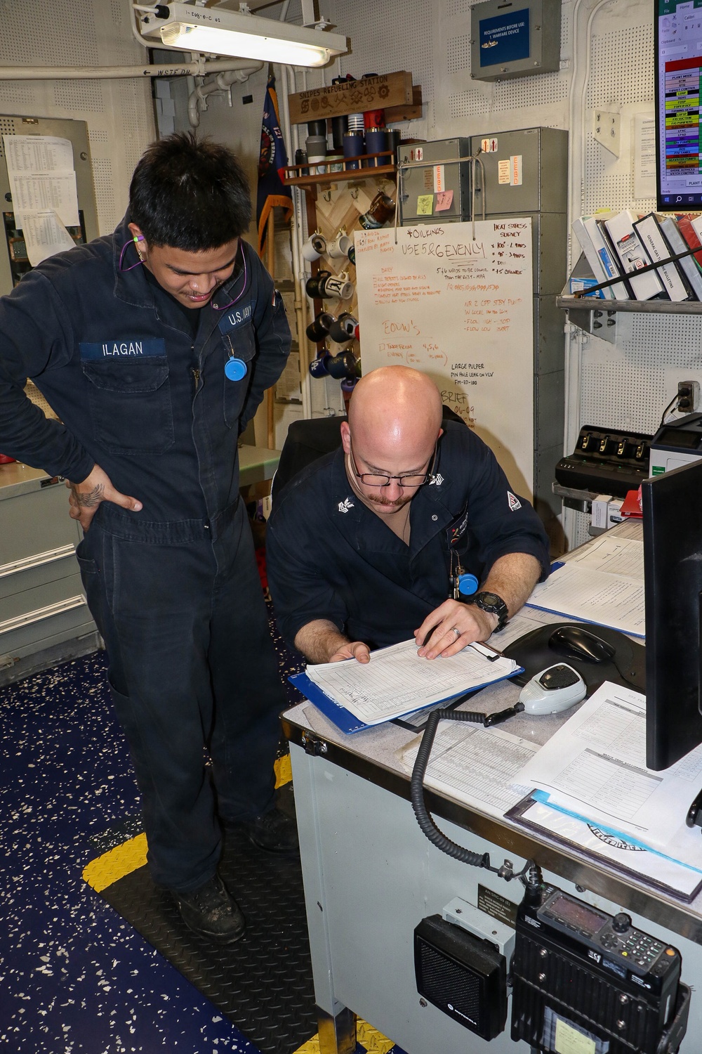 Two U.S. Navy sailors in dark blue coveralls are reviewing paperwork at a workstation aboard a naval vessel. The sailor on the left, identified by his name tape as 'ILAGAN,' is standing and observing while a bald sailor wearing glasses is seated and writing or reviewing documents on a clipboard. The workspace includes a whiteboard with operational notes, radio equipment, and various administrative materials typical of a ship's engineering or damage control station.