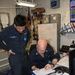 Two U.S. Navy sailors in dark blue coveralls are reviewing paperwork at a workstation aboard a naval vessel. The sailor on the left, identified by his name tape as 'ILAGAN,' is standing and observing while a bald sailor wearing glasses is seated and writing or reviewing documents on a clipboard. The workspace includes a whiteboard with operational notes, radio equipment, and various administrative materials typical of a ship's engineering or damage control station.