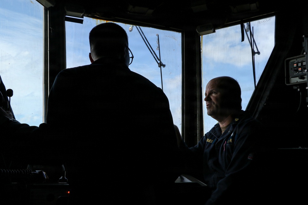 Two individuals are inside what appears to be a ship's bridge or wheelhouse, with one person seated and wearing a dark naval or maritime uniform with a name tag, and another person standing with their back to the camera. The scene is heavily backlit by windows showing a blue sky with clouds and exterior rigging or mast structures. The high-contrast lighting creates strong silhouettes, particularly of the standing figure.