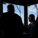 Two individuals are inside what appears to be a ship's bridge or wheelhouse, with one person seated and wearing a dark naval or maritime uniform with a name tag, and another person standing with their back to the camera. The scene is heavily backlit by windows showing a blue sky with clouds and exterior rigging or mast structures. The high-contrast lighting creates strong silhouettes, particularly of the standing figure.