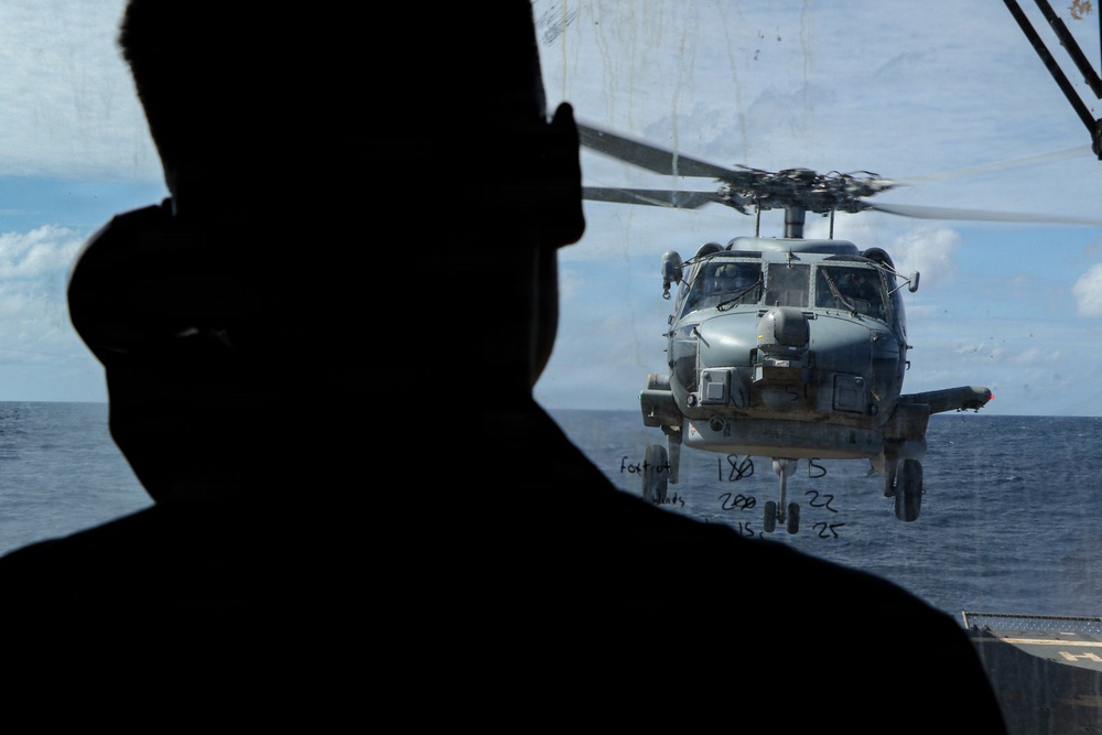 A silhouetted figure, likely military personnel, observes a naval helicopter hovering near a ship's deck over open ocean. The helicopter appears to be an MH-60 Seahawk variant, positioned close to the vessel, suggesting a deck landing or hover operation. The image is taken from inside or near a ship's bridge or observation area, with handwritten numerical notations visible on the glass.