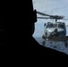A silhouetted figure, likely military personnel, observes a naval helicopter hovering near a ship's deck over open ocean. The helicopter appears to be an MH-60 Seahawk variant, positioned close to the vessel, suggesting a deck landing or hover operation. The image is taken from inside or near a ship's bridge or observation area, with handwritten numerical notations visible on the glass.