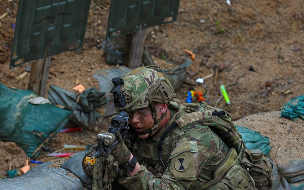 A U.S. Army soldier in MultiCam (OCP) camouflage uniform is positioned in a fortified fighting position surrounded by sandbags, aiming a rifle with an optic sight. The soldier wears a combat helmet with accessories and a unit patch visible on the left shoulder, consistent with the 10th Mountain Division insignia. The setting appears to be a military training exercise based on the presence of colored markers and training aids scattered in the background.