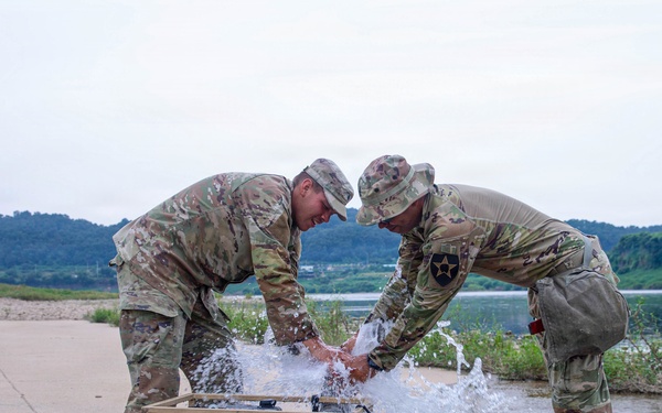 Two U.S. Army soldiers in OCP (Operational Camouflage Pattern) uniforms are working together to manage a water pump that appears to be leaking or malfunctioning, with water spraying forcefully from a connection point. The scene is set near a flooded or waterlogged area with a river and green hills visible in the background. One soldier displays a 2nd Infantry Division patch on their left shoulder.
