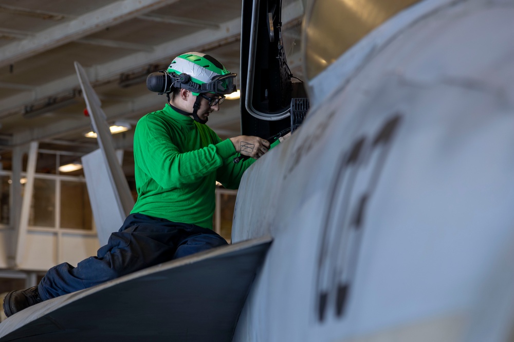A naval aviation technician wearing a green jersey and helmet with ear protection is performing maintenance or inspection work on a military aircraft, likely aboard an aircraft carrier hangar deck. The green jersey indicates the individual is likely a catapult and arresting gear crew member or aviation ordnanceman. The setting appears to be an enclosed hangar bay with overhead lighting and structural elements visible in the background.