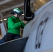A naval aviation technician wearing a green jersey and helmet with ear protection is performing maintenance or inspection work on a military aircraft, likely aboard an aircraft carrier hangar deck. The green jersey indicates the individual is likely a catapult and arresting gear crew member or aviation ordnanceman. The setting appears to be an enclosed hangar bay with overhead lighting and structural elements visible in the background.