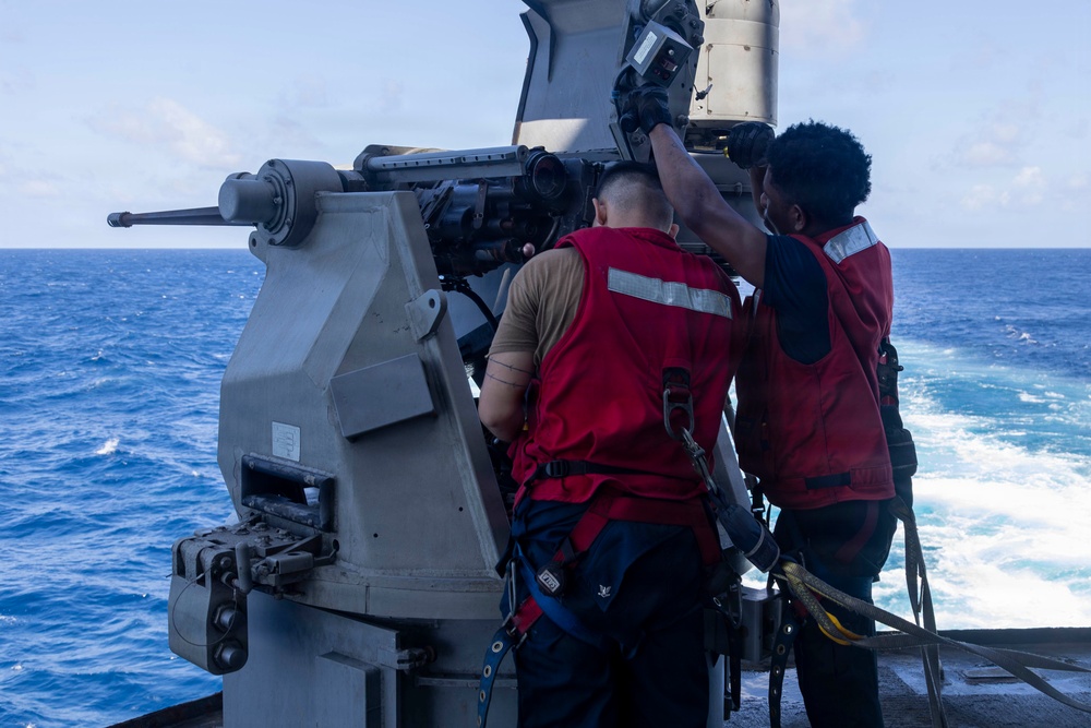 Two naval personnel wearing red safety vests and harnesses are working on a deck-mounted naval gun system aboard a warship underway at sea. The crew members appear to be conducting maintenance or inspection of what appears to be a Mk 38 machine gun system or similar naval deck gun. The ship is moving through open blue ocean waters, visible by the wake trailing behind.
