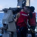 Two naval personnel wearing red safety vests and harnesses are working on a deck-mounted naval gun system aboard a warship underway at sea. The crew members appear to be conducting maintenance or inspection of what appears to be a Mk 38 machine gun system or similar naval deck gun. The ship is moving through open blue ocean waters, visible by the wake trailing behind.