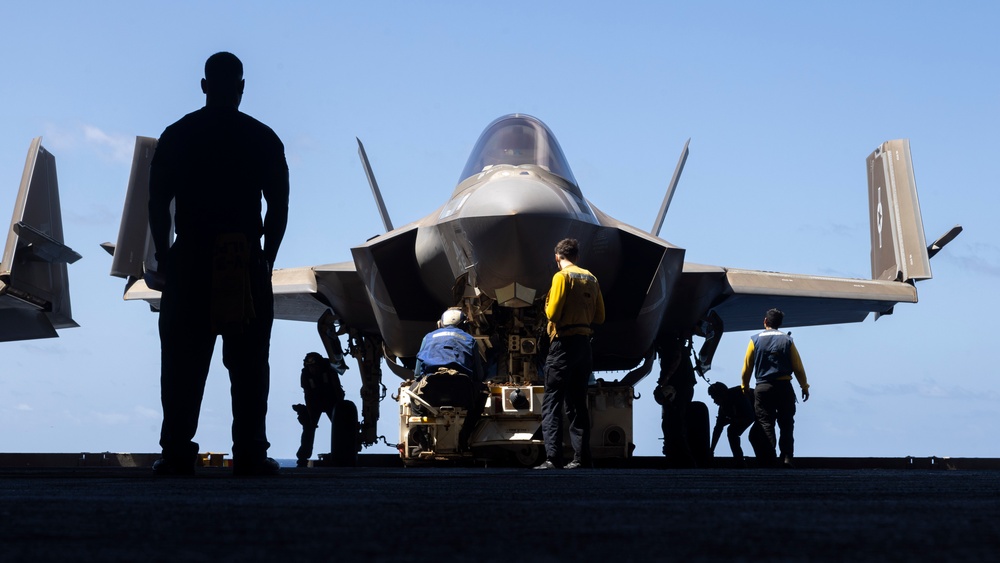 A military jet aircraft, consistent with an F-35 variant, is positioned on what appears to be an aircraft carrier flight deck. Several ground crew personnel in various colored jerseys are conducting pre-flight or maintenance operations around the aircraft. The image is shot in strong backlighting, creating dramatic silhouettes against a clear blue sky.
