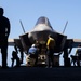 A military jet aircraft, consistent with an F-35 variant, is positioned on what appears to be an aircraft carrier flight deck. Several ground crew personnel in various colored jerseys are conducting pre-flight or maintenance operations around the aircraft. The image is shot in strong backlighting, creating dramatic silhouettes against a clear blue sky.