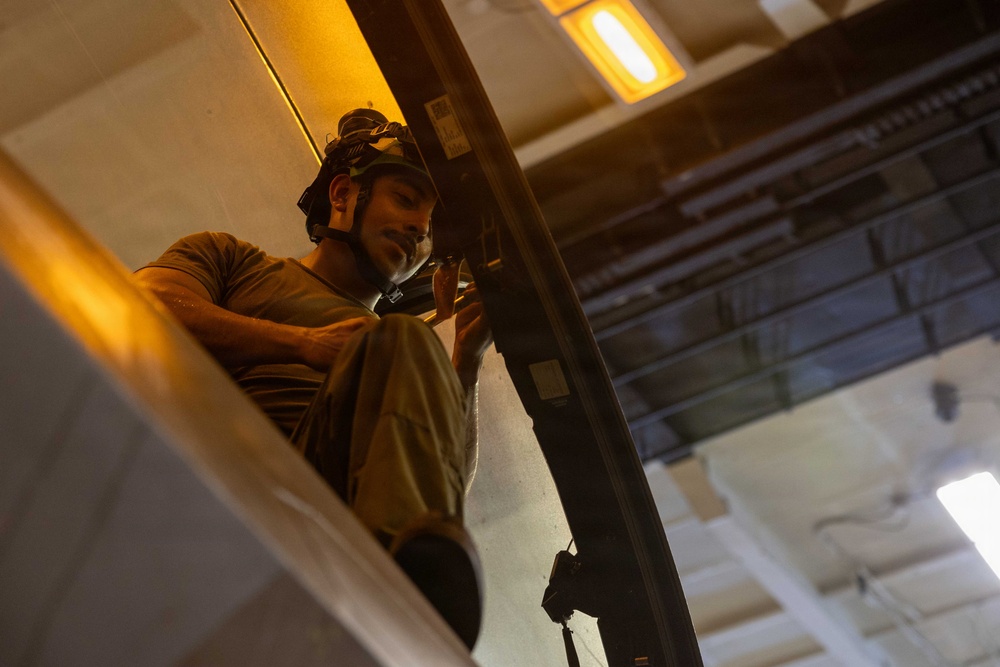 A bearded male worker wearing a headlamp is photographed from a low angle while working at an elevated position, likely on a ladder or elevated platform inside an industrial or commercial building. He appears to be performing maintenance or installation work on a structural beam or frame. The warm orange lighting from nearby fixtures contrasts with the cooler ambient light of the facility.