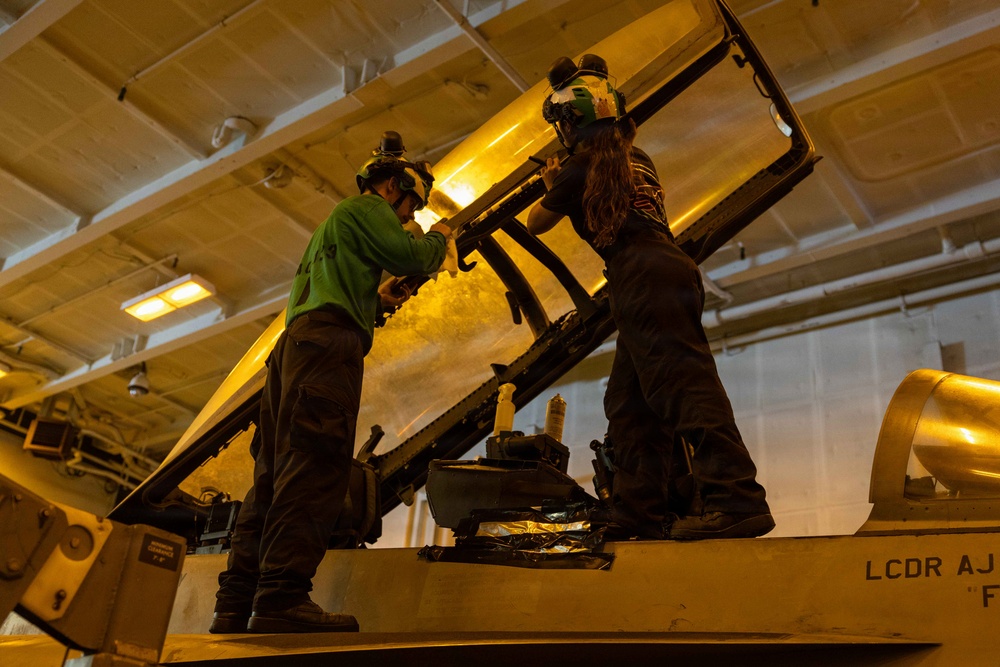 Two maintenance personnel are working on what appears to be a military aircraft cockpit canopy in an enclosed hangar bay, likely aboard a naval vessel. They are standing on top of the aircraft and appear to be performing maintenance or inspection on the canopy frame. A partial name stencil reading 'LCDR AJ' is visible on the aircraft fuselage.