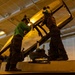 Two maintenance personnel are working on what appears to be a military aircraft cockpit canopy in an enclosed hangar bay, likely aboard a naval vessel. They are standing on top of the aircraft and appear to be performing maintenance or inspection on the canopy frame. A partial name stencil reading 'LCDR AJ' is visible on the aircraft fuselage.