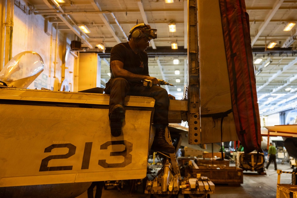 A maintenance technician is seated on top of a military aircraft marked with the number '213', working with a power tool in what appears to be a naval vessel's hangar bay. The worker is wearing hearing protection and safety goggles pushed up on his helmet while performing maintenance on the aircraft's wing or fuselage area. The large indoor hangar contains multiple aircraft and personnel visible in the background.