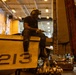A maintenance technician is seated on top of a military aircraft marked with the number '213', working with a power tool in what appears to be a naval vessel's hangar bay. The worker is wearing hearing protection and safety goggles pushed up on his helmet while performing maintenance on the aircraft's wing or fuselage area. The large indoor hangar contains multiple aircraft and personnel visible in the background.