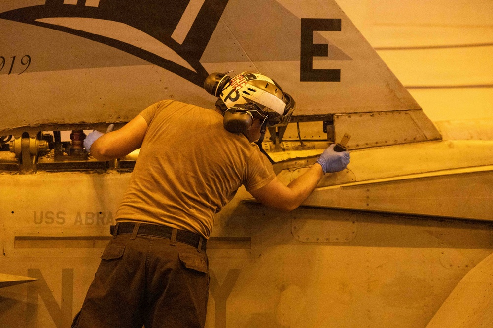 A military technician or aviation mechanic is performing maintenance work on a naval aircraft, likely in a hangar bay. The worker is wearing protective gear including hearing protection, goggles, and blue latex gloves while working on the aircraft's fuselage or wing junction area. The partial text 'USS ABRA' is visible on the aircraft, indicating this is aboard a U.S. Navy vessel, likely the USS Abraham Lincoln.