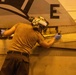 A military technician or aviation mechanic is performing maintenance work on a naval aircraft, likely in a hangar bay. The worker is wearing protective gear including hearing protection, goggles, and blue latex gloves while working on the aircraft's fuselage or wing junction area. The partial text 'USS ABRA' is visible on the aircraft, indicating this is aboard a U.S. Navy vessel, likely the USS Abraham Lincoln.