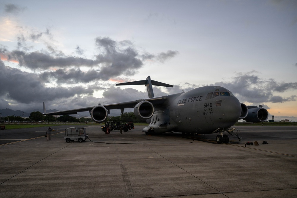 A U.S. Air Force C-17 Globemaster III military transport aircraft, tail number 5146, is parked on a tarmac at dusk or dawn. The aircraft bears markings indicating assignment to the 15th Wing and 154th Wing. Ground crew personnel and support equipment are visible around the aircraft, suggesting pre-flight or post-flight servicing operations.