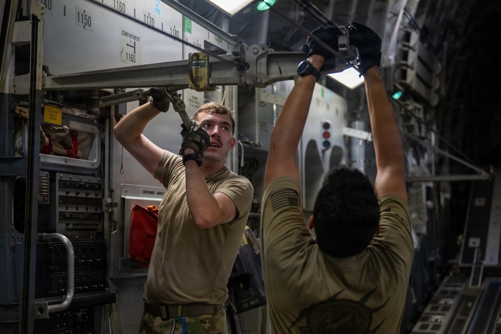 Two military personnel in olive drab uniforms and tactical gloves are working inside what appears to be a large military transport aircraft, likely a C-17 Globemaster III. They are manipulating overhead cargo tie-down or locking mechanisms along the interior fuselage rail system. The interior shows numbered station markings (1140-1180) and various aircraft systems panels.
