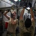 Two military personnel in olive drab uniforms and tactical gloves are working inside what appears to be a large military transport aircraft, likely a C-17 Globemaster III. They are manipulating overhead cargo tie-down or locking mechanisms along the interior fuselage rail system. The interior shows numbered station markings (1140-1180) and various aircraft systems panels.