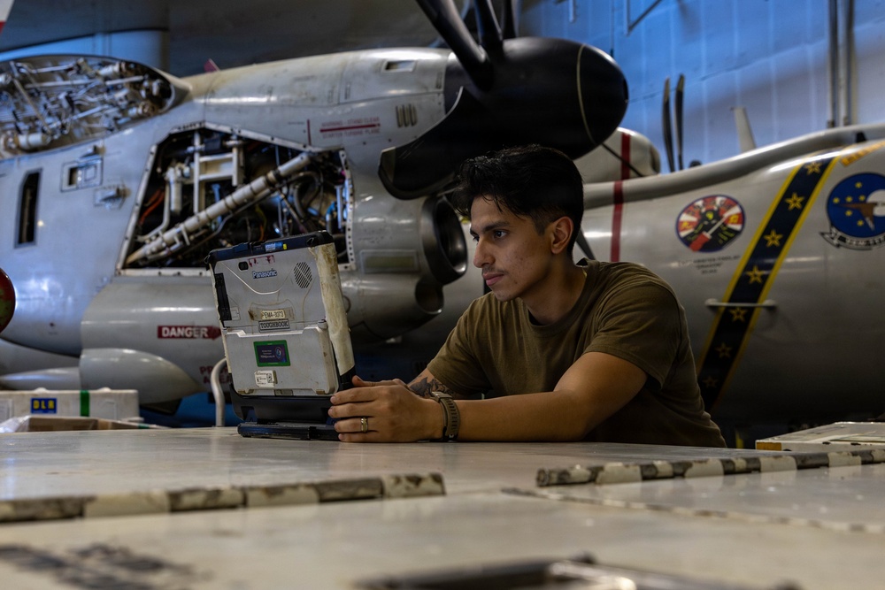 A military technician or mechanic is working in an aircraft hangar, using a ruggedized Panasonic Toughbook laptop on a work surface. Behind him, a large military propeller aircraft (appears to be a C-2 or similar turboprop) is undergoing maintenance with its engine cowling open. The setting is a military aviation maintenance facility.