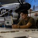 A military technician or mechanic is working in an aircraft hangar, using a ruggedized Panasonic Toughbook laptop on a work surface. Behind him, a large military propeller aircraft (appears to be a C-2 or similar turboprop) is undergoing maintenance with its engine cowling open. The setting is a military aviation maintenance facility.