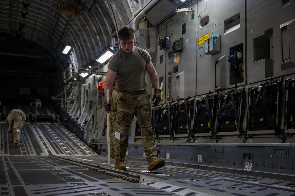 A military service member in MultiCam/OCP camouflage uniform is working inside the cargo hold of a large military transport aircraft, likely a C-17 Globemaster III. The individual appears to be preparing or configuring the cargo floor, holding what appears to be a tie-down bar or cargo restraint tool. A second service member is visible in the background near the rear of the aircraft.
