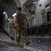 A military service member in MultiCam/OCP camouflage uniform is working inside the cargo hold of a large military transport aircraft, likely a C-17 Globemaster III. The individual appears to be preparing or configuring the cargo floor, holding what appears to be a tie-down bar or cargo restraint tool. A second service member is visible in the background near the rear of the aircraft.
