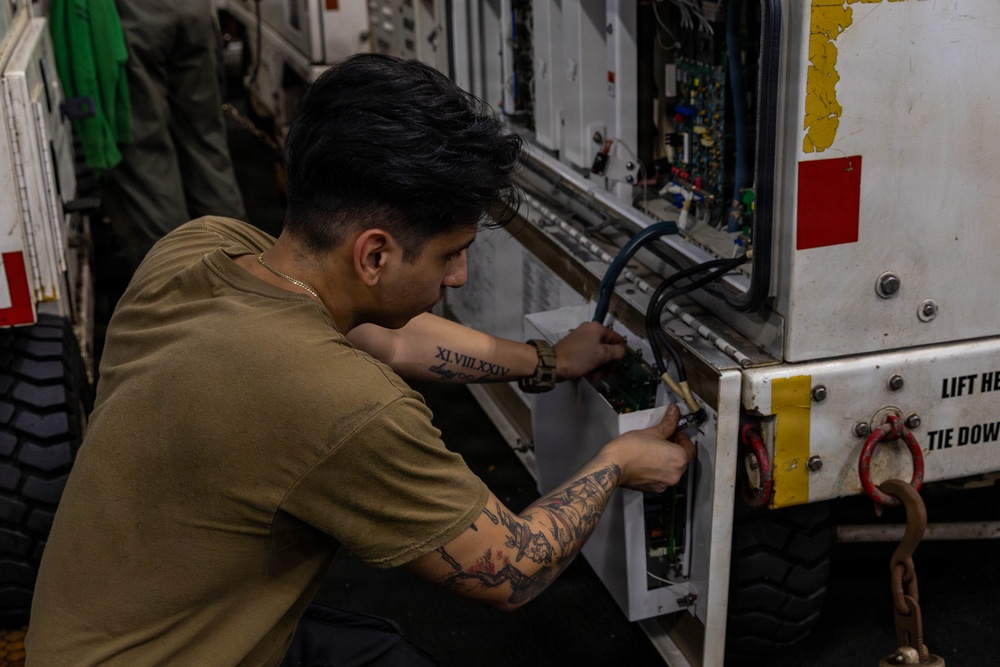 A young male technician in an olive/tan t-shirt is performing maintenance or repair work on an open electrical or electronic panel of a military or industrial vehicle. He is crouching and using tools to work on wiring and circuit components inside the panel. The equipment bears markings including 'LIFT H' and 'TIE DOW' (partially visible), suggesting military ground support equipment.