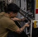 A young male technician in an olive/tan t-shirt is performing maintenance or repair work on an open electrical or electronic panel of a military or industrial vehicle. He is crouching and using tools to work on wiring and circuit components inside the panel. The equipment bears markings including 'LIFT H' and 'TIE DOW' (partially visible), suggesting military ground support equipment.